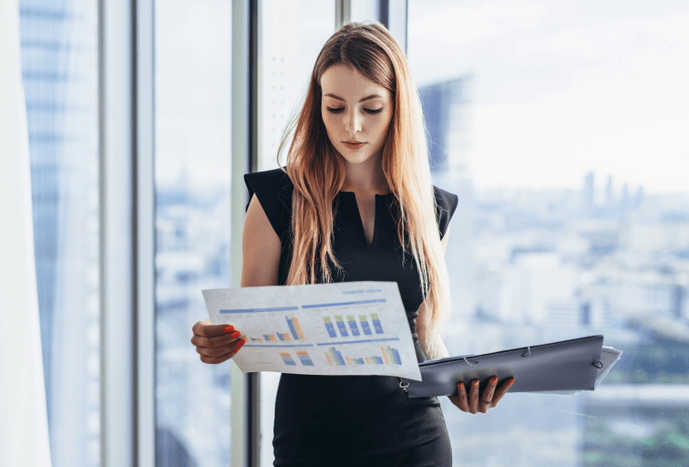 Female financial analyst holding papers studying documents standing against window with city view.